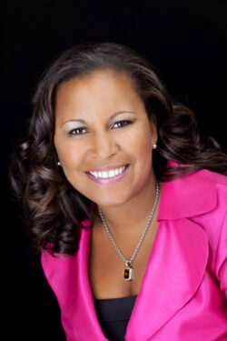 Vibrant professional headshot of a smiling woman in a bright pink blazer and amber pendant necklace, photographed against a black background by Exposures Photography in Toronto.