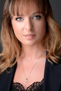 Warmly lit professional lifestyle headshot of a woman with blonde hair and bangs, wearing a black blazer and lace top, by Exposures Photography